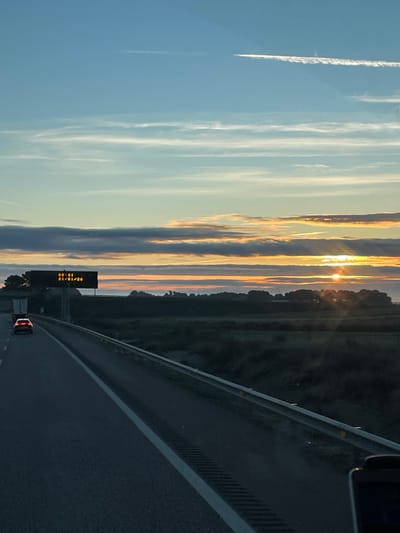 A highway stretches towards the horizon, reflecting twilight light. The sun's rays peek through vertical cloud formations against a blue sky. A distant road sign is visible. A guardrail flanks the road. The perspective suggests the photo was taken from a moving vehicle.