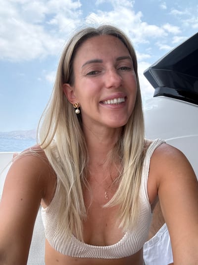 A woman with blonde hair smiles at the camera while on a boat. She is wearing a white bikini top and a necklace with a small pendant. The woman is outdoors, with a cloudy sky and the sea visible in the background. This image was taken in Agia Marina, Greece.