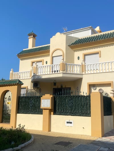 The image shows a light yellow, two-story house with a dark green tiled roof and white balustrades on a balcony. The house features an arched window on the upper floor, a chimney with a green decorative cap, and a white satellite dish on the roof. A dark green wrought-iron fence encloses the property, with an arched gate on the left side and an orange pillar on the right. A beige electrical box is mounted on a pillar in front of the fence. The ground in front of the house is paved with light brown and pink square tiles. A small, overgrown garden with a curved concrete border is visible in the foreground on the left. The sky is clear and blue. The scene is located in Cancelada, Spain.