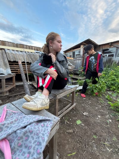 This image depicts two individuals in an outdoor setting that appears to be a rural or dilapidated area, possibly in Маркидоновка, Russia. The scene is characterized by weathered wooden structures, overgrown vegetation, and a cloudy sky, suggesting it might be daytime with overcast weather.

In the foreground, a young person, likely female given the long hair and earrings, is seated on a wooden structure. She is wearing a black leather jacket, black pants with red and white stripes, and light-colored sneakers. Her gaze is directed upwards and to the right, and her posture suggests a relaxed or contemplative mood.

Behind and above her, another individual, likely male, is standing. He is wearing a black baseball cap, a dark jacket over a graphic t-shirt with a bird or owl design, and black pants. He is also wearing red slip-on shoes. He is looking downwards towards the ground.

The overall impression is one of a casual, perhaps slightly neglected, outdoor environment. The wooden structures could be part of an old building or fence. The presence of a rake leaning against a wooden beam suggests some form of outdoor activity or maintenance. The image captures a moment of pause or interaction between the two individuals in this rustic location.