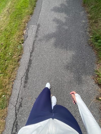The image displays a high-angle, first-person perspective of an individual walking on a paved pathway in Skanderborg, Denmark. The person is dressed in dark blue athletic leggings and white athletic shoes. A light-colored, long-sleeved athletic top, featuring a zippered front and a thumbhole on the left sleeve, is visible from the waist down, with the left hand partially protruding through the thumbhole. The pathway is constructed of dark gray asphalt, showing visible cracks and signs of wear, particularly along its left edge. Strips of green grass, interspersed with dry patches and weeds, border both sides of the path. Shadows, indicative of an overhead light source, are cast upon the walking surface.