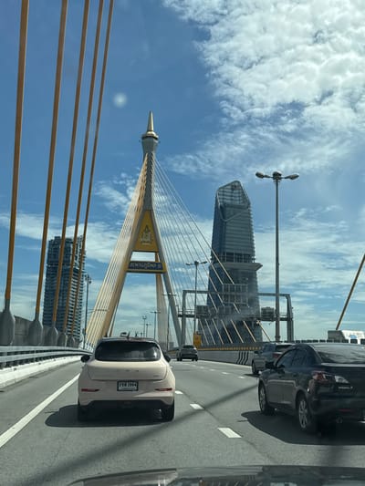 The image captures a view from a vehicle driving on a multi-lane bridge in Song Khanong Subdistrict, Thailand. The foreground shows the rear of a light-colored car with a Thai license plate reading "3ชร 2964." To the right, a black sedan with "Mazda 3" visible on its trunk is in the adjacent lane. In the midground and background, the bridge's distinctive yellow support cables are prominent, leading to a tall, slender pylon adorned with a crest and Thai text. Beyond the bridge, a modern, irregularly shaped skyscraper with a glass facade stands against a bright blue sky with scattered white clouds. Another, taller, gray skyscraper is visible to the left of the bridge structure. Streetlights are present along the bridge, and the overall scene suggests a bright, daytime environment.