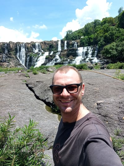 A man wearing sunglasses and a dark t-shirt smiles at the camera in front of a multi-tiered waterfall in a rocky landscape. Green foliage is visible in the foreground and on the forested hills behind the waterfall. The sky is blue with scattered white clouds. This is the Datanla waterfall in the Da Lat region, approximately 15 kilometers from the city of Da Lat, in the Lâm Đồng Province of Vietnam, not Tân Phú.