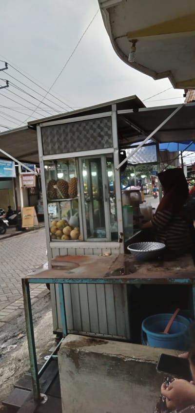 A street food stall operates on a paved street in Tanjungan, Indonesia, under an overcast sky. The primary subject is a metal-framed stall with glass panels displaying pineapples and a cluster of yellow fruits, likely mangoes or guavas. A person, partially obscured, wears a striped top and a dark head covering, standing behind the stall next to a counter with a silver textured bowl and various containers. Below the counter, a blue bucket containing an orange scoop is visible. In the immediate foreground, a person's hands hold a dark-colored smartphone. The ground around the stall is a mix of concrete, dirt, and some standing liquid. In the background, a commercial street features other buildings, an awning, and a sign displaying "BEBEK TANJUNGAN" (Tanjungan Duck). Several motorcycles are parked along the street, and other individuals are visible further down the road, including one on a motorcycle. Overhead, multiple electrical wires crisscross, and a spiral fluorescent light bulb hangs from an adjacent structure.