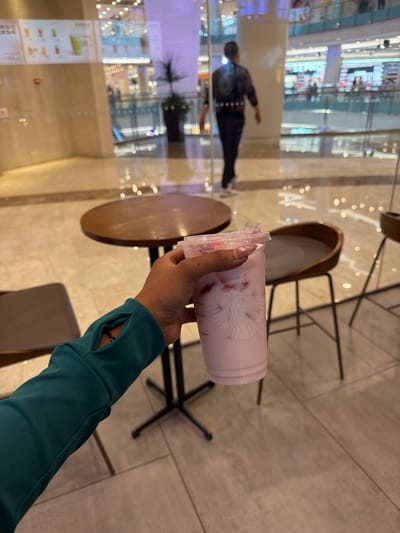 This image captures a first-person perspective shot within a modern indoor commercial space, likely a shopping mall or large complex in Yuhuatai District, China. The central focus is a hand holding a Starbucks cup filled with a light pink beverage containing visible fruit pieces, possibly strawberries. The person's hand has light-colored, possibly artificial, fingernails, and they are wearing a teal/dark green long-sleeved top with a thumbhole, revealing a black beaded bracelet or watch on their wrist. The Starbucks siren logo is clearly visible on the cup.
The background features a reflective glass wall or window, creating numerous bokeh light reflections and mirroring the interior environment. Through the reflections, one can discern blurry figures of people, including one person walking away from the camera, dressed in dark pants and a patterned top. Also visible in the reflections are parts of store displays, blurred architectural elements, and what appears to be a menu board with some unreadable text.
The setting includes brown wooden round tables and chairs with black metal frames, suggesting a cafe or seating area. The floor is a light-colored, possibly tiled surface with faint grid patterns. The space is brightly illuminated by artificial lighting, indicating an indoor environment where the time of day is not distinctly natural. The overall impression is that of someone taking a break to enjoy a drink in a bustling commercial hub.