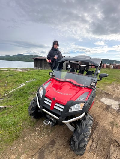 A woman stands next to a red ATV in Bolshaya Rechka, Russia. The ATV has a black roof and seats. The woman is holding a glass. In the background, there is a wooden building, a body of water, and trees on rolling hills. The sky is overcast with clouds. The ground is a mix of dirt and grass, with some puddles visible.