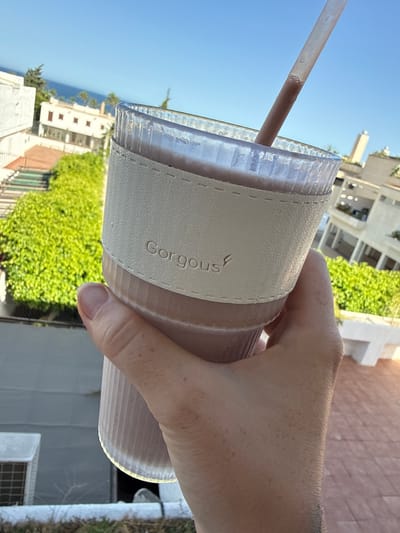 A person's hand holds a translucent, ribbed plastic cup with a textured, light-colored sleeve. The sleeve has stitched detailing and the word "Gorgous" embossed on it. A light brown straw extends from the cup into the clear liquid inside. The background shows a bright blue sky, green trees, and white buildings with balconies, suggesting an outdoor setting. The lighting indicates it is daytime, likely sunny given the clear sky and bright illumination. The scene appears relaxed and casual, possibly a person enjoying a drink outdoors in a warm climate. The location is identified as Aguadulce, Spain, which aligns with the architectural style and vegetation visible.