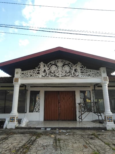 The image displays the front facade of a single-story building located in Cijedil, Indonesia. The structure features a red-brown pitched roof and a white exterior. A central entrance consists of two closed, brown corrugated metal roll-up doors. Dark-tinted windows flank the doors on either side. Above the entrance, a white, ornate, scrolled decorative panel spans horizontally. Two white, fluted columns with decorative capitals and colorful tiled bases support an overhang on the facade. The ground immediately in front of the building is a cracked concrete or asphalt surface with sparse vegetative growth. White plastic bags and minor debris are visible near the base of the left column. A wooden, multi-sectioned structure leans against the building's facade to the right of the entrance. The sky overhead is blue with scattered white clouds, and multiple black utility lines traverse the upper portion of the frame. No individuals are present.