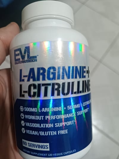 An indoor scene in Parkent, Uzbekistan, depicts a close-up view of a white plastic supplement bottle held by a human left hand. The bottle is capped with a white screw-on lid. Its label, featuring a holographic finish with rainbow reflections, prominently displays "EVL EVLUTIONNUTRITION" and large blue text reading "L-ARGININE + L-CITRULLINE". Further details on the label include "500MG L-ARGININE + 500MG L-CITRULINE", "WORKOUT PERFORMANCE SUPPORT", "VASODILATION SUPPORT", and "VEGAN/GLUTEN FREE". The label also specifies "60 SERVINGS" and "DIETARY SUPPLEMENT 120 VEGGIE CAPSULES". The thumb and fingers of the individual's left hand are visible on the left side of the frame, gripping the bottle. The background consists of a light-colored, possibly white or light grey, tiled surface with discernible diagonal grout lines.