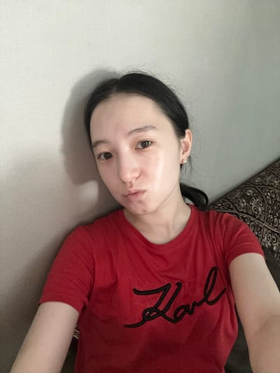 A young woman with dark hair tied back and fair skin with visible pores and a few blemishes is shown from the chest up. She is wearing a red t-shirt with a black, script-style logo that appears to say "Karl." She is looking directly at the camera with her lips slightly pursed and has a pearl earring in her right ear. She is leaning against a textured, light-colored wall that casts a shadow to her left. To the right, a patterned textile, possibly part of a sofa, is visible. The lighting suggests an indoor setting. The image was taken in Nookat, Kyrgyzstan.