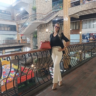 A woman in a black blazer and white pants stands on a balcony overlooking a multi-level indoor space. She wears sunglasses and a watch, and a red purse rests on the railing beside her. The building features stone walls, multiple levels with ornate railings, and various shops. The floor is tiled with a herringbone pattern. A colorful play area is visible below the balcony.