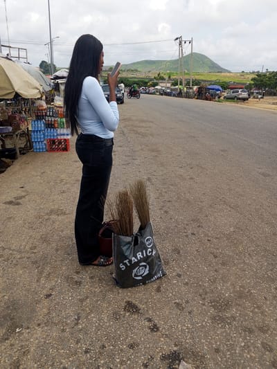 A young woman with long black hair, wearing a light blue long-sleeved shirt and dark wash wide-leg jeans, stands on a paved road, holding a pink smartphone. To her left, on the side of the road, is a market stall with various items, including crates of bottled drinks. A large black tote bag filled with brooms rests on the ground near the woman's feet. The background shows a road with passing vehicles, including motorcycles and cars, and a distant green mountain under a cloudy sky. The scene appears to be a roadside market in Pasere, Nigeria.
