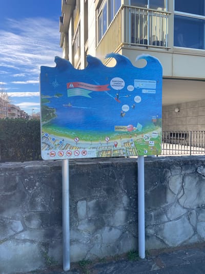 An outdoor informational sign, shaped like ocean waves, stands prominently in a city environment in Zarautz, Spain. The sign is mounted on two silver metallic poles embedded in the ground, positioned directly in front of a weathered stone wall.

The sign features a detailed, colorful map and diagram on a blue background, illustrating coastal activities and regulations. The upper portion depicts a section of the ocean with a compass rose, a red motorboat moving leftwards, and several individuals engaged in water sports such as surfing and swimming. Text in Basque is visible, including a prominent warning triangle with "ERNE KORRONTEEKIN" (Beware of currents) and "ZAINZARIK GABEKO HONDARTZA" (Unsupervised Beach). Another section indicates "URAREN KALITATEA BIKAINA" (Excellent Water Quality).

The lower part of the sign transitions to a green-and-tan depiction of a coastline, featuring a detailed map of the beach, "HONDARTZA," and adjacent urban areas, identifying "Santa Barbara." Numerous circular red prohibition symbols with diagonal lines are displayed across the beach area, indicating restrictions such as no littering, no dogs, no fires, and other regulations.

In the background, a multi-story building with light-colored facades and numerous windows is visible. The building features a prominent balcony with a light-colored railing on one of its upper floors. To the far left, beyond some low green hedges, other urban structures are partially visible. The sky is bright blue with scattered wispy cirrus clouds. The scene is illuminated by natural daylight, suggesting a clear day. No individuals are directly visible in the immediate vicinity of the sign or in the background.