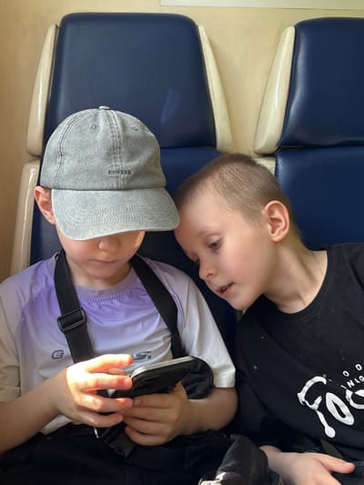 In Vyatskiye Polyany, Russia, two young boys are seated side-by-side in what appears to be a public transport vehicle, indicated by the blue upholstered seats with light-colored, possibly cream, plastic dividers. The boy on the left wears a grey denim-textured baseball cap with "NOFTAN" embroidered on the front, a purple and white long-sleeved athletic shirt with a stylized 'E' or 'G' logo on the chest, and dark lower garments with a black shoulder strap. He is holding a dark-colored smartphone with both hands, looking down at its screen, with direct light illuminating his hands. The boy on the right, with short light-brown hair, wears a black long-sleeved t-shirt with white graphic text, including "OOD" and "Foos" (partially visible, likely part of "FOOD"), and a thin silver chain around his neck. He is leaning towards the boy on the left, intently observing the smartphone screen. The background consists of similar blue seats and a light-colored wall.