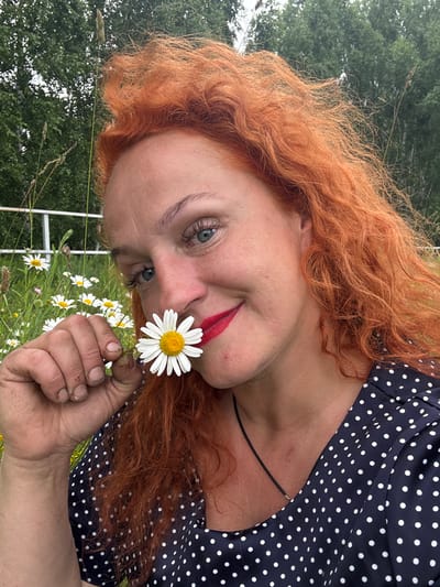 A woman with long, wavy, bright red hair and blue eyes is featured in this outdoor portrait. She is wearing a navy blue dress with white polka dots. Her face is partially obscured as she leans in towards the camera, holding a white daisy with a yellow center in her right hand. She has red lipstick on, and her expression appears playful or curious. The background consists of greenery, likely a field or garden, with more daisies visible. The lighting suggests it is daytime, possibly overcast given the lack of strong shadows. The scene evokes a sense of nature and simplicity. The provided location context is Privolzhsky, Russia.
