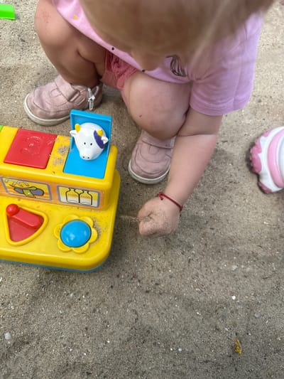 A child in a pink outfit and pink shoes is playing with a yellow toy in a sandpit. The toy has buttons and images of farm animals and milk bottles. The child's hands and legs are covered in sand. The scene appears to be outdoors, likely in a park or playground, given the presence of a sandpit. The context of Donchovci, Bulgaria, suggests this is a local scene.