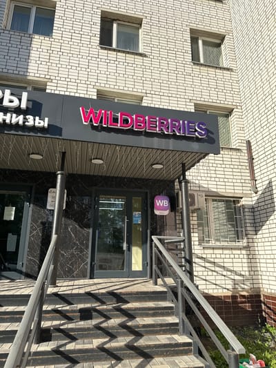 The image shows the entrance to a Wildberries store in the city of Rubtsovsk, Russia. The store is located on the ground floor of a white brick building. The entrance features a dark marble facade with a prominent "WILDERRIES" sign in pink and purple lettering. To the left of the entrance, a portion of another sign is visible, with white letters on a dark background. A set of concrete stairs with metal railings leads up to the entrance. A small white sign with a purple "WB" logo is mounted on the facade to the left of the door. An air conditioning unit is visible on the wall above the entrance. The scene is illuminated by sunlight, casting strong shadows on the stairs and the building's facade. There are no people visible in the image.