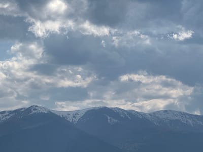 The image depicts a mountain range with snow-capped peaks under a cloudy sky. The mountains are dark blue-grey and appear to be in the distance. The clouds are a mix of light and dark grey. Based on the prompt, the location is Bokilovtsi, Bulgaria.
