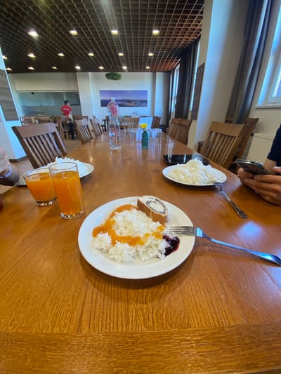 The image shows a dining table in Loučná nad Desnou, Czechia. The table is long and wooden, with several wooden chairs arranged along its sides. On the table, there are plates of food: one with rice, jam, and a pastry; another with rice. There are also two glasses of orange juice and several clear glasses. Two forks are visible near the plates. A person is holding a mobile phone above the table, and another person's hand with a bracelet is visible at the edge of the frame. The background shows a window, curtains, and a view into another part of the building.