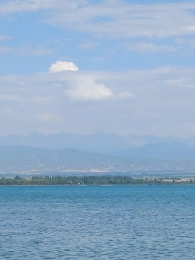 A serene landscape photograph captures a wide body of blue water in the foreground, its surface rippled by gentle waves. In the midground, a lush green tree line forms a stark contrast against the pale blue sky. Beyond the trees, distant mountains fade into a hazy blue, their peaks softened by atmospheric perspective. The sky is dotted with soft white clouds, one particularly prominent cumulus formation floating in the upper left quadrant. The scene is tranquil and evokes a sense of natural beauty. The location is identified as Kirpichny, Kyrgyzstan.