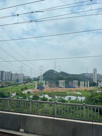 The image, taken in Dongguan, China, captures a construction scene alongside a body of water and a line of high-voltage power towers. In the foreground, a concrete railing with an ornamental pattern separates the viewer from the vista. Beyond the railing, the landscape unfolds with lush green vegetation bordering a calm body of water.
The middle ground is dominated by a significant infrastructure project. Multiple large concrete pillars, part of what appears to be an elevated highway or bridge, are under construction. Scaffolding and construction materials are visible around these structures. The power towers, characterized by their intricate lattice designs, march in a line across the scene, signifying the presence of significant electrical infrastructure.
In the background, a range of buildings, including high-rise apartment blocks and other structures, are nestled amongst a backdrop of densely wooded hills. The sky is overcast with scattered clouds, suggesting a day with diffused light, possibly early morning or late afternoon. The overall impression is one of development and the juxtaposition of urban expansion with natural elements and existing infrastructure. No people are clearly visible in the image.