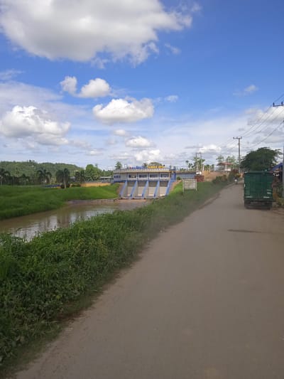 The image depicts a dirt road leading towards a dam structure labeled "JAMBO AYER" in the city of Langkahan, Indonesia. The sky is blue with scattered white clouds. To the left of the road is a body of water and a lush green grassy area, with palm trees and a forested hill in the background. The dam is a concrete structure with blue accents and a white building on top. To the right of the road, there are utility poles with electrical wires, a green truck, and a motorcycle. The road surface is unpaved and appears to be made of dirt and gravel. The vegetation along the roadside is dense and green.