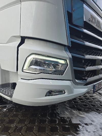 The image displays a close-up, angled view of the lower-right front section of a white DAF heavy goods vehicle. The vehicle is situated on a dark grey permeable paver surface, which is wet and covered in white soap suds, indicating it is undergoing or has recently completed a wash in Ostend, Belgium.

The right front of the truck features a prominent white bumper and fender. The right headlight unit is illuminated, showing distinct LED daytime running lights. Below the main headlight, a smaller, oval-shaped fog light is integrated into the bumper. To the right, a section of the truck's black grille, characterized by a honeycomb pattern, is visible, along with a partial view of the "DAF" logo. A blue Belgian license plate, partially visible at the bottom right of the grille, displays "2-GCK-". On the far left, a black, textured step-plate for cab access is present. Water droplets and white soap suds are actively dripping from the vehicle's underside onto the wet paver ground. No individuals are present in the frame.