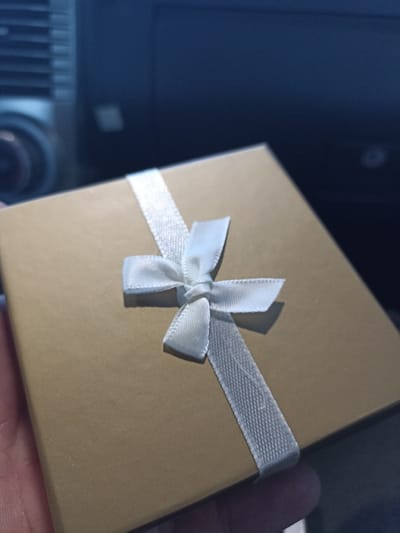 A square, gold-colored gift box is tied with a white ribbon. The ribbon diagonally crosses the box and is tied in a bow at the center. The box appears to be held in someone's hand, with a dark vehicle dashboard visible in the background. The lighting is dim, casting shadows on the box and ribbon.