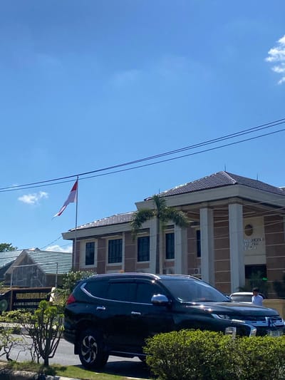 The image shows a clear, bright day in a city in Jawa, Indonesia. The foreground is dominated by the side and front of a black SUV parked on the side of a road, with lush green bushes surrounding it and a section of manicured lawn. Behind the SUV, a building with a tiled roof and cream-colored columns stands prominently. Above the building, an Indonesian flag, red and white, flutters on a flagpole against a bright blue sky with a few scattered white clouds. To the left of the main building, another building with a similar tiled roof is visible, and a sign with Indonesian text is mounted on a fence. In the background, another building and some trees can be seen. A person in a white shirt is visible near the entrance of the main building. The overall scene suggests an official or government building located in a well-maintained urban environment.