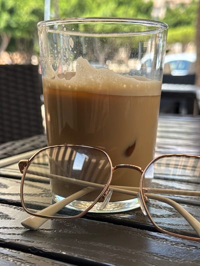 A close-up, eye-level, outdoor shot captures a glass of iced coffee with a frothy top and ice cubes resting on a dark, weathered wooden table. In the foreground, a pair of rose gold-rimmed glasses are placed partially over the coffee glass, with their arms resting on the table. The background is softly blurred, showing green foliage and what appears to be a white structure, possibly a building. The setting suggests a cafe or outdoor dining area. The location is identified as Campillo Del Moro, Spain. There are no people visible in the image.