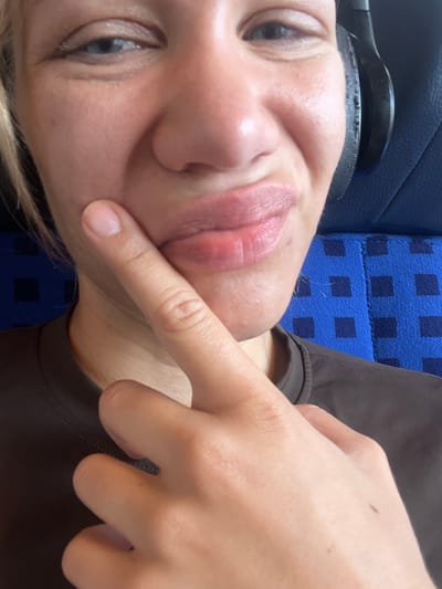 A close-up, head-and-shoulders portrait of a fair-skinned person, likely female, is captured. The subject is wearing a dark brown t-shirt and has blonde hair. A pair of black headphones is visible over their right ear. The individual is touching their cheek with their right index finger, positioned near their lips. The person's lips appear slightly swollen and have a rosy tint. The background consists of dark blue seats with a checkered pattern, typical of public transportation. The lighting is somewhat diffused, creating soft shadows on the face. The location is identified as Ampfing, Germany.