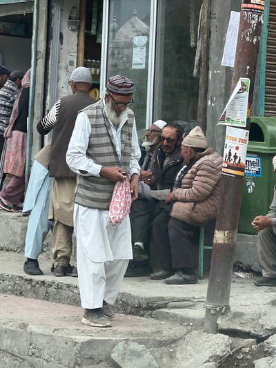 The image depicts a street scene in Dras, India, with several men gathered near a shop and a utility pole. The central figure, a man wearing a white shirt, a checkered vest, and white pants, walks towards the viewer, holding a pink and white patterned cloth bag in his right hand. He has a white beard and wears glasses and a striped cap. To his left, another man in a brown vest with striped sleeves and a white cap is partially visible. Further to the left, a woman in a pink headscarf and a light blue shalwar kameez is seen walking away.
To the right of the central figure, a group of men are seated. One man in a light brown puffer jacket and a beige cap is looking down and to the left. Next to him, a man wearing sunglasses and a dark jacket is looking towards the viewer. Another man with a white cap and beard is seated further back.
The environment includes a storefront with a glass window, a wooden pillar, and a metal utility pole adorned with posters. A green trash bin is visible behind the seated men. The ground is uneven, with visible cracks in the concrete. The lighting suggests an overcast day.