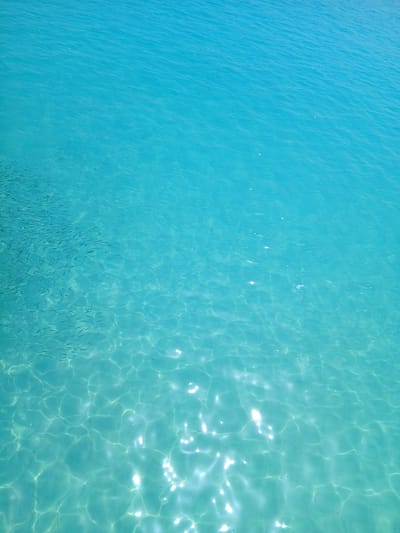 The image depicts a top-down view of clear, turquoise seawater, likely in Sidi Abdel Rahman, Egypt. Sunlight penetrates the water, creating bright, shimmering patterns on the surface. A large school of small fish is visible swimming close to the bottom. The water varies in color, ranging from a deeper, solid turquoise on the left to a lighter, more translucent shade on the right where the sun penetrates more strongly. The surface of the water is mostly smooth except for some small ripples and the sun reflecting from the waves.