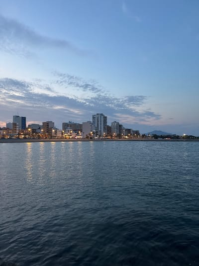 A tranquil coastal scene at dusk or early evening in Vinaròs, Spain, is captured in this wide-angle view. The image presents a horizontal composition of the sky, a bustling cityscape, and the calm sea.

The upper portion of the frame features a twilight sky, transitioning from a lighter blue near the horizon to a deeper indigo overhead. Wispy, elongated clouds, illuminated by the fading daylight, stretch across the sky.

Along the horizon, a vibrant cityscape unfolds, showcasing numerous multi-story buildings, likely residential or commercial, lining the beachfront. Many windows and streetlights are illuminated, casting a warm yellow and orange glow across the scene, indicating the time of day. In the far distance, a faint, dark mountain range is visible against the fading light.

The lower half of the image is dominated by the calm, dark blue waters of the sea. Gentle ripples animate the surface, and the illuminated city lights create shimmering, elongated reflections that stretch across the water, adding a dynamic sparkle to the serene expanse.

The scene evokes a peaceful and picturesque atmosphere, capturing the quiet beauty of a Mediterranean town as day transitions into night. There is no visible text in the image.