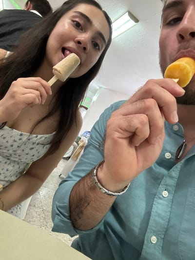 Two people are eating popsicles in what appears to be an indoor setting. A woman in the foreground is holding and licking a beige-colored popsicle. She has long, dark hair and is wearing a floral-patterned top. A man is partially visible in the background, also eating a popsicle. He's wearing a blue button-down shirt and a silver bracelet. The lighting is bright and even, suggesting artificial illumination. The background is out of focus, showing tables and chairs, and there's what appears to be a window with green and white light shining through. The setting seems to be a casual eatery or similar space. I cannot confirm the location in Chipilo de Francisco Javier Mina (Chipilo), Mexico with certainty based on the image alone.