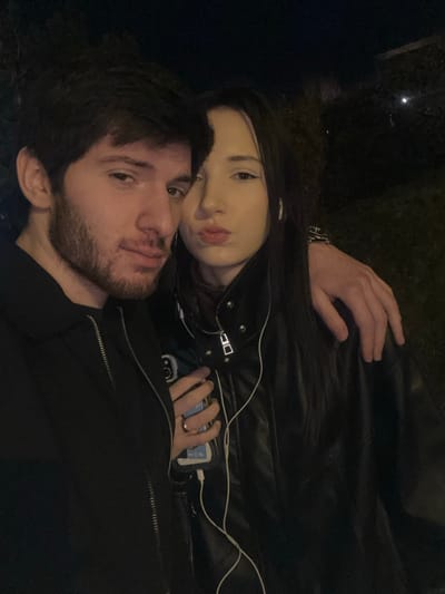 A young couple poses closely together at night. The man has dark hair and a beard. The woman, with long dark hair, is behind him. They both look at the camera. The woman wears a black leather jacket, and a hand with a watch is visible above her. A phone plugged into a charger is in her other hand. The man wears a black jacket. The lighting is dim, giving the scene a nighttime aesthetic.
