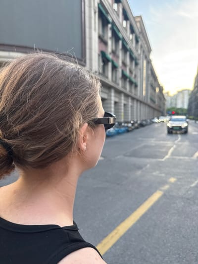 This portrait-oriented image, presented rotated ninety degrees clockwise, captures a person looking skyward in an urban setting. In the foreground, seen from behind, is the head and neck of a person with light brown hair, likely styled in a low ponytail or bun. Sunglasses are perched on top of their head, and they are wearing a black sleeveless top. A silver earring is visible in their ear.

The person's gaze is directed upwards towards a towering building that dominates the left side of the frame (when viewed upright). This multi-story structure features numerous windows and distinctive green or teal accents, extending high into a bright, clear sky, suggesting daytime. On the right side of the frame, a paved city street stretches into the distance, marked by a prominent yellow dividing line. A line of parked bicycles or scooters is visible along the street, and further down, several cars, including one with a visible red brake light or traffic signal, indicate ongoing activity. The bright sunlight suggests the time of day is likely late morning or afternoon. The scene depicts a casual moment of observation within Putuo District, China, characterized by its urban architecture and street life. No discernible text appears in the image.