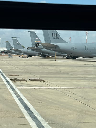 The image depicts a tarmac scene with multiple large military transport aircraft parked in a row, viewed from a slightly elevated, angled perspective, likely from within a vehicle or building. The aircraft are gray and appear to be KC-135 Stratotankers, identified by their distinctive tail fins and markings. The tail of one aircraft clearly shows "HH" and the number "8880," along with an Air Force star insignia. Another tail in the foreground shows "HH" and "03576." Further down the line, a tail displays "AFRC" and "714SG." Two figures, one in a yellow shirt and another in darker clothing, are visible near the wing of one aircraft, seemingly engaged in maintenance or inspection work, with a portable ladder structure nearby. The tarmac is paved concrete with visible markings, including a prominent double white line with a dark stripe between them running diagonally across the foreground. In the background, two tall poles with red and white markings are visible, likely airport navigation lights or beacons, and some distant buildings and structures suggest an airfield environment. The sky above is a hazy light blue with some clouds. The overall scene is one of an active, though perhaps not immediately busy, military airfield. The image does not provide any specific visual cues to confirm the location in the city of Yagel, Israel.