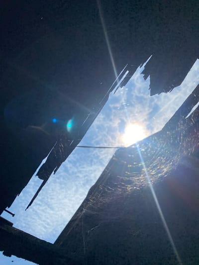 The image captures a view looking upwards through a damaged roof, revealing a bright sky with scattered clouds. The sun is a prominent feature, casting strong lens flares and illuminating a delicate spiderweb strung between the broken roof materials. The roof itself appears to be made of dark, possibly charred or weathered material, with irregular, jagged edges. The scene suggests an abandoned or damaged structure, possibly in a less developed area, though no specific landmarks or people are visible to definitively place it within Banjarmasin, Indonesia. The overall impression is one of decay and natural elements reclaiming a man-made space.