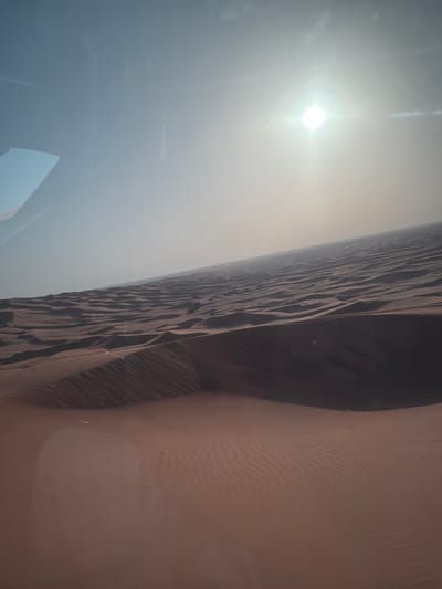 The image depicts a sunlit desert landscape. The scene is dominated by a large sand dune, with visible ripples and textures. A hazy atmosphere obscures the background, with the sun appearing as a bright orb in the upper left portion of the image. The color palette consists of earth tones, ranging from light beige to dark brown. The environment suggests the dunes of Al Madam in the United Arab Emirates.
