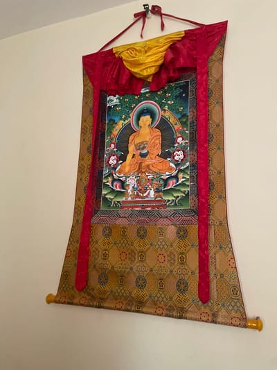 A Thangka, a traditional Tibetan Buddhist painting, is prominently displayed, hanging vertically against a plain, light-colored wall. The central artwork within the Thangka depicts a seated Buddha figure, rendered in warm orange and golden tones. The Buddha is shown in a meditative posture, with one hand resting in the lap and the other gently touching the earth, a gesture commonly associated with the Bhumisparsha Mudra. A radiant halo and an aureole encircle the Buddha's head and body. The background of the painting features lush green foliage, possibly a landscape with trees bearing fruits or flowers, and hints of a blue sky.
The painting is mounted on a larger textile scroll made of a rich, golden-brown brocade fabric. This fabric is intricately patterned with geometric and stylized floral motifs in shades of blue, red, and gold, creating a luxurious frame for the central image. Vibrant red silk borders frame the painting on all sides, extending upwards and downwards. At the top of the Thangka, draped over the red borders, are folds of bright red and golden-yellow silk, forming decorative elements. The entire Thangka is suspended from a horizontal wooden dowel at the top, and a similar dowel, with yellow finials, is partially visible at the bottom, providing weight and structure. No activity is taking place, and no visible text is present in the image.