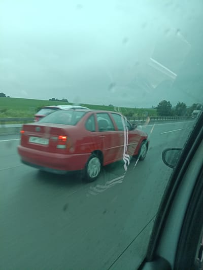A red sedan is driving on a highway in Divišov, Czechia, on a cloudy day. The car is red with a license plate that is unreadable due to motion blur. There is another car behind it that is white. The highway has multiple lanes and is bordered by a grassy field and trees on the right. The image is taken from inside another vehicle, as evidenced by the reflection on the windshield. The weather appears to be rainy or at least humid, with water droplets visible on the windshield.