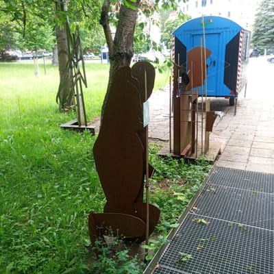 The image depicts an outdoor art installation in Eisenach, Germany, with a focus on rusty metal sculptures. In the foreground, a large, dark brown, abstract metal figure stands on the left, partially obscuring a metal frame structure. To the right of the abstract figure, a blue trailer with the text "Kunststation Oepfershausen" is visible, suggesting it is part of an art station or exhibition. The trailer has black and red-striped detailing and a blue door. Behind the trailer, more abstract metal elements and structures are present. The ground is a paved walkway with scattered debris, adjacent to a grassy area with trees and foliage. A metal grate covers a part of the walkway in the lower right corner. The scene appears to be in a public space, possibly a park or an urban area with green spaces.