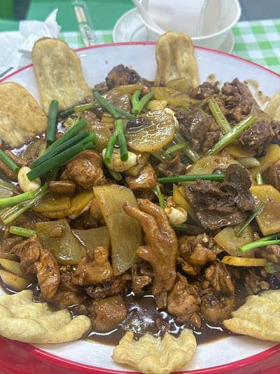 This image depicts a large, prepared meal served on a checkered tablecloth in Licheng District, China. The central subject is a substantial, shallow, white serving dish with a prominent red rim, filled with a saucy chicken and potato preparation. The dish contains numerous irregular pieces of cooked chicken, including a distinct brown chicken foot located centrally. Interspersed among the chicken are thin, light-brown slices of potato and vibrant green chopped scallions. All ingredients are coated in a dark brown, glossy sauce. Several pieces of light-golden fried dough are arranged around the perimeter of the dish, some flat and oblong, others with a ruffled or crimped edge, partially submerged in the sauce. In the blurred background, a green and white checkered tablecloth is visible, along with a white ceramic cup and saucer, and chopsticks in a green and white wrapper. A white fork is partially visible to the left. No people are present in the scene.