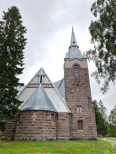 The image shows a stone church with a tall, pointed steeple, surrounded by trees. The church features a combination of light-colored stucco and rough-hewn stone construction. The steeple is made of gray metal and has several small windows. The lower portion of the church and the base of the steeple are constructed from large, reddish-brown stones. The church has several windows, some with lattice work. In the foreground, there is green grass. The sky is overcast. The provided information indicates the location is the city of Melnikovo, Russia.