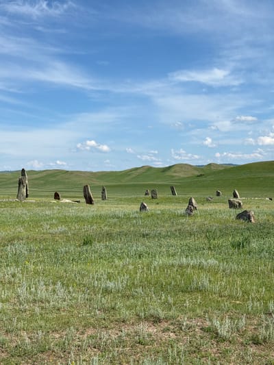 The image depicts a landscape scene in the vicinity of Safronov, Russia. The foreground is dominated by a vast, grassy plain with scattered wildflowers and dry patches of earth. In the midground, a collection of ancient standing stones, or menhirs, are arranged in a field. These stones vary in size and shape, with some standing tall and others lying on the ground. Beyond the stones, rolling green hills form the background, under a bright blue sky dotted with white, wispy clouds. The overall atmosphere is one of ancient history and natural beauty.