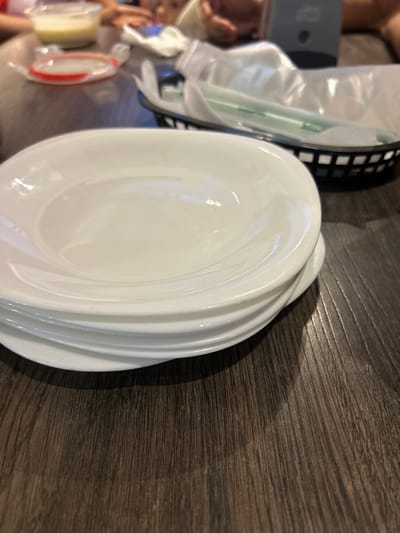 A stack of white, ridged dinner plates sits on a dark wooden surface. A black plastic basket containing a wrapped utensil is nearby. A doll or figurine can be partially seen alongside a small plastic container. The composition features a strong contrast between the smooth white plates and the textured wood.