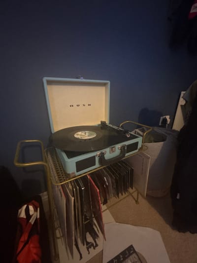The image displays an indoor scene, likely a personal living space in Newton Upon Derwent, United Kingdom, under dim lighting, suggesting evening or a dimly lit room. A light blue, portable record player, designed in a retro briefcase style, is open with a vinyl record resting on its platter. The brand name "BUSH" is visible in black, vertically aligned text on the cream-colored inside surface of the open lid.

To the right of the record player, a gold-colored metal rack holds a collection of vinyl records, neatly stacked upright. Various record sleeves, including red, white, and dark-colored ones, are visible within the rack. The record player and rack are situated on a light-colored, possibly beige or light brown, carpeted floor. Additional record sleeves or papers are scattered on the floor to the right. A light-colored woven storage basket or container is partially visible behind the record setup. In the bottom right corner of the frame, a red and white fabric item, possibly a bag or clothing, can be seen. The background on the left is a dark blue wall. No people are present in the image.
