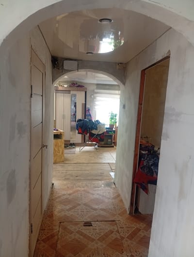 The image depicts an interior hallway with an arched doorway leading into a room. The hallway walls are unfinished, showing exposed plaster and some patchy paint. A light-colored wooden door is visible on the left, with a silver doorknob. The floor is a combination of wooden planks and patterned tiles.

In the room beyond the archway, a wardrobe with mirrored doors is present, along with a clothes rack holding various garments, predominantly dark blue and red. A small wooden stand is also visible. Further in the background, a window with blinds allows natural light to enter, illuminating a potted plant on a shelf.

On the right side of the hallway, there's an open doorway leading to another area. This area appears cluttered, with items stacked and covered by fabric, including a prominent red textile. The ceiling in the hallway is reflective, showing a distorted view of lights and possibly other objects.

The overall impression is of an unfinished or in-progress renovation within a residential property in Khomutovo, Russia, characterized by a blend of raw and completed construction elements. No people are visible in the image.