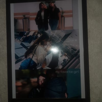 The image displays a framed collage of three photographs. The top photograph shows two young women posing for a selfie. The middle photograph is a reflection in a mirror, capturing a woman taking a selfie. The bottom photograph shows two people embracing, with the text "my favorite girl" written on the right side. The collage is mounted on a textured, grey wall, and the overall setting appears to be indoors. There is no information in the image to suggest the location is in the city of Қосшы, Kazakhstan.